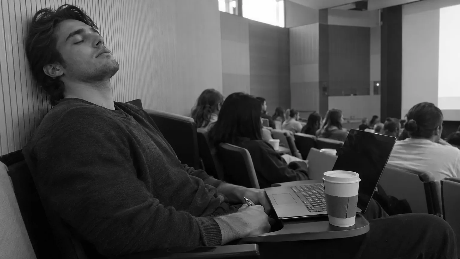 A university student asleep mid-lecture, slumped back in his seat with his head tipped back and a faint smile, a pen still in his hand, other students around him facing the front.